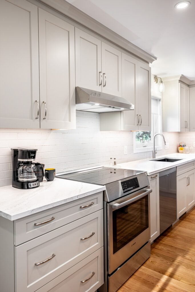 Modern kitchen with white cabinets, stainless steel oven, dishwasher, coffee maker, and a sink under a window. White subway tile backsplash and wooden floor are also visible. Bright, clean, and well-lit space.
