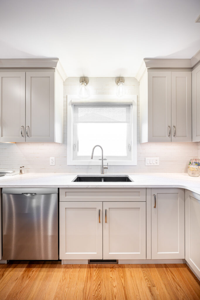 Modern kitchen with light gray cabinets, a stainless steel dishwasher, a large window above a sink with a faucet, white countertops, and wood flooring under bright lighting.