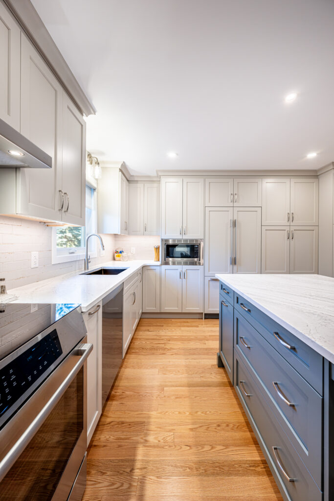 A modern kitchen with light gray cabinets, stainless steel appliances, a large island with marble countertops, wooden floors, a built-in microwave, and under-cabinet lighting.