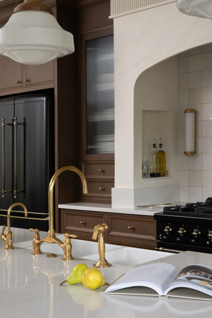 A kitchen with a white countertop, gold faucets, an open cookbook, two pears, a black stove, and dark wood cabinets. Olive oil bottles sit in a nook by cream-colored tiled walls.