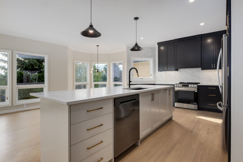 Modern kitchen with a large island, white countertops, black cabinets, stainless steel appliances, pendant lights, and multiple large windows letting in natural light.