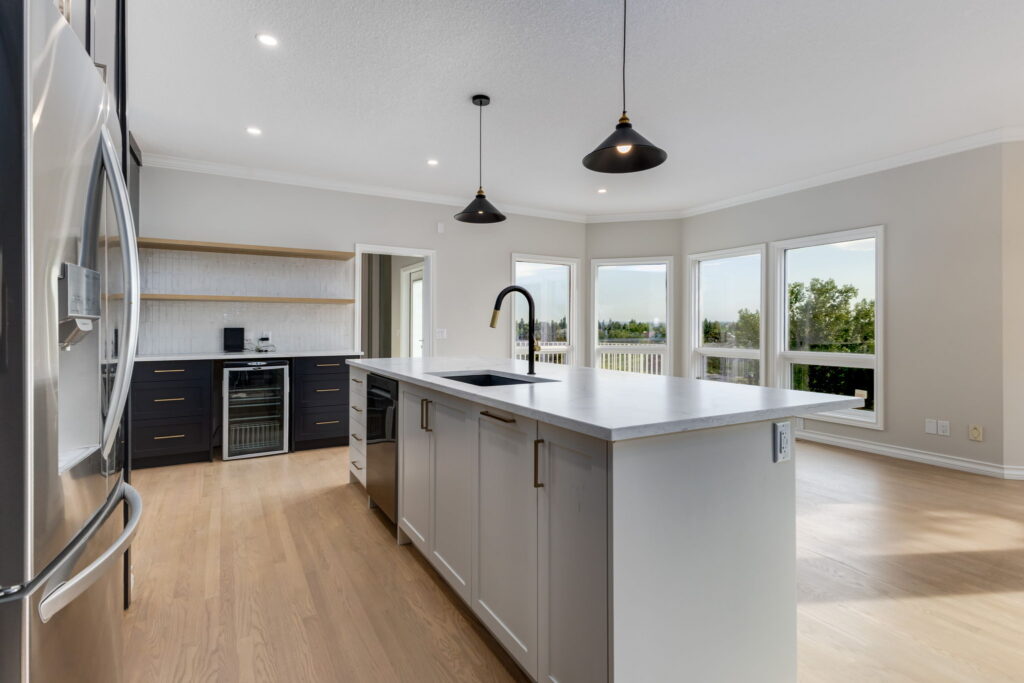 Modern kitchen with a large island, sink, pendant lights, stainless steel appliances, dark lower cabinets, open shelves, and large windows overlooking a scenic outdoor view.
