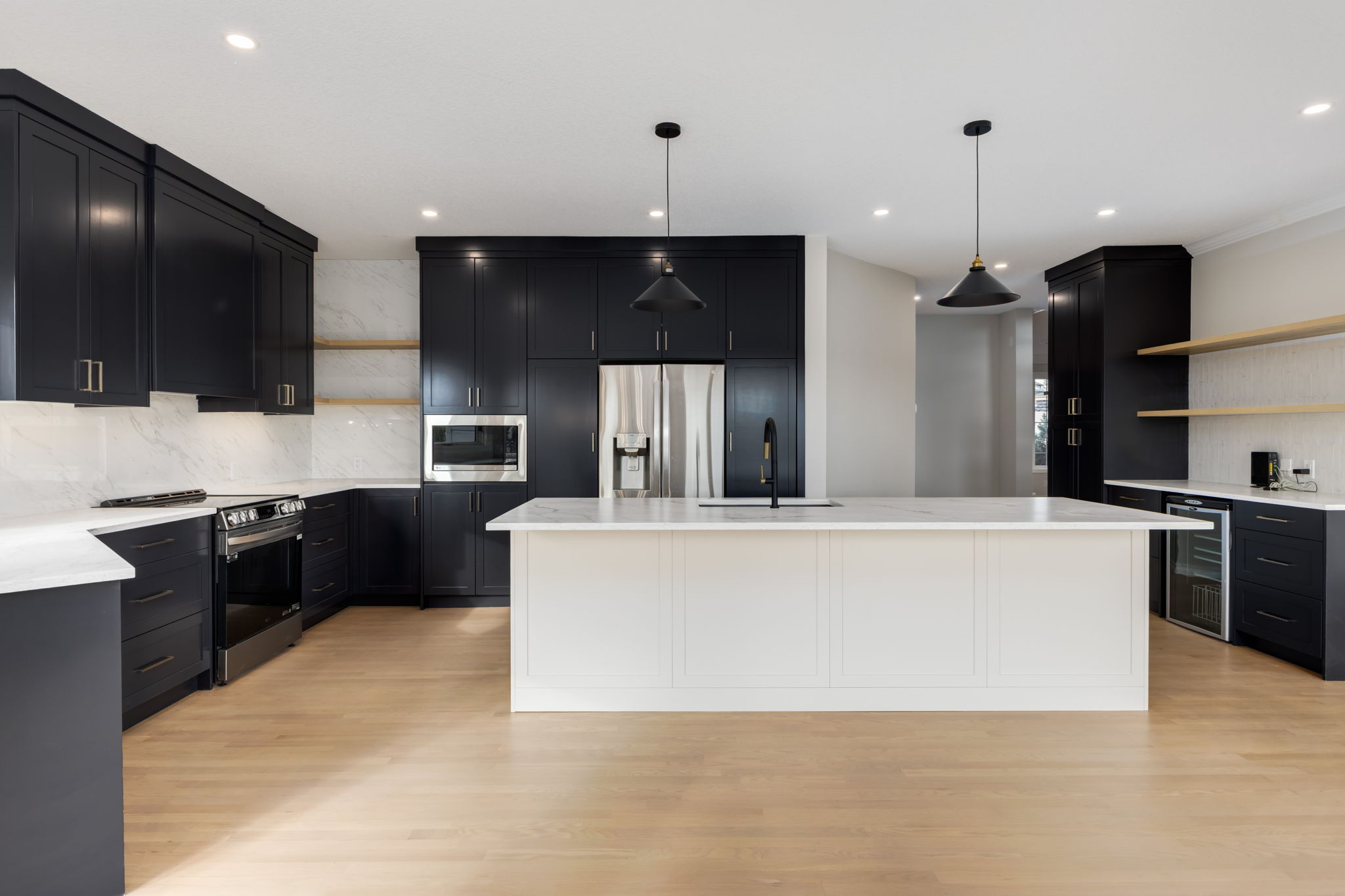 Modern kitchen with black cabinets, white countertops, a large white island, stainless steel appliances, pendant lights, open wooden shelves, and light wood flooring. The space is bright and minimalist.