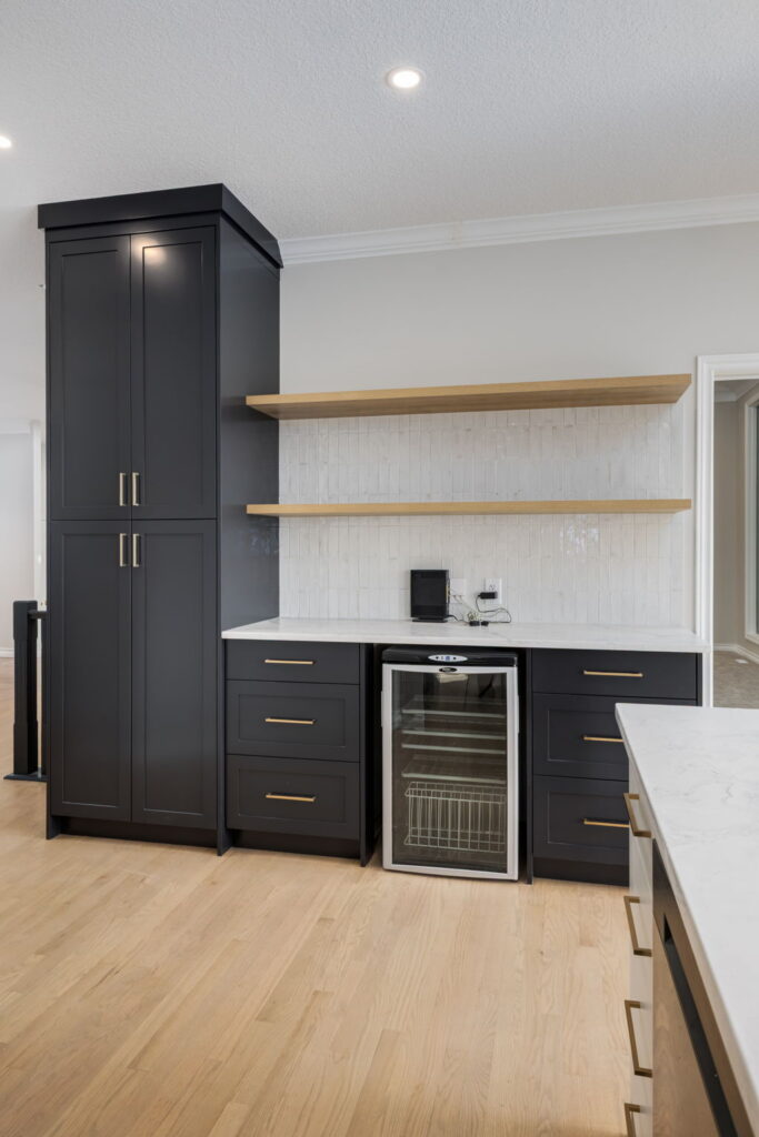 Modern kitchen with black cabinets, wooden shelves, white backsplash, light wood floor, and a small glass-front refrigerator beneath the counter. Countertop has a black speaker and small kitchen accessories.