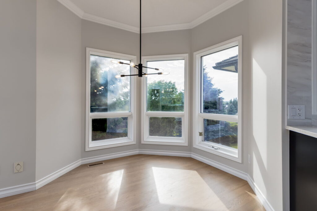 Bright, empty breakfast nook with large bay windows, modern chandelier, light wood flooring, and white walls, overlooking a green outdoor landscape.