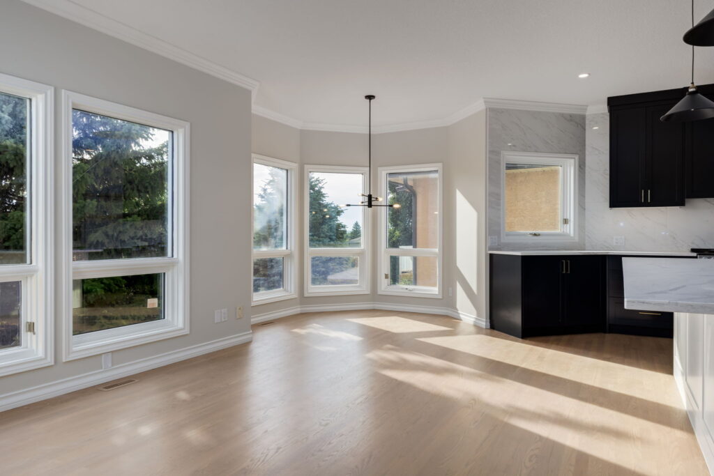 Bright, empty dining area with large bay windows, light wood floors, and an adjacent kitchen with black cabinets and marble backsplash. Sunlight streams in, providing a clear view of trees outside.
