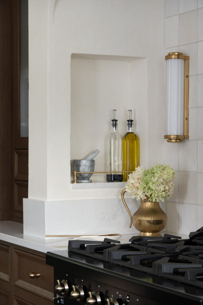 A kitchen nook with a marble mortar and pestle, two glass oil dispensers, a brass pitcher holding white flowers, and a stove in the foreground. The wall is white with a brass wall sconce and tiled backsplash.