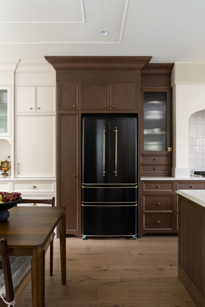 A modern kitchen featuring a black refrigerator with gold handles, surrounded by dark wood cabinets, a wood dining table with chairs, and light-colored countertops and walls.