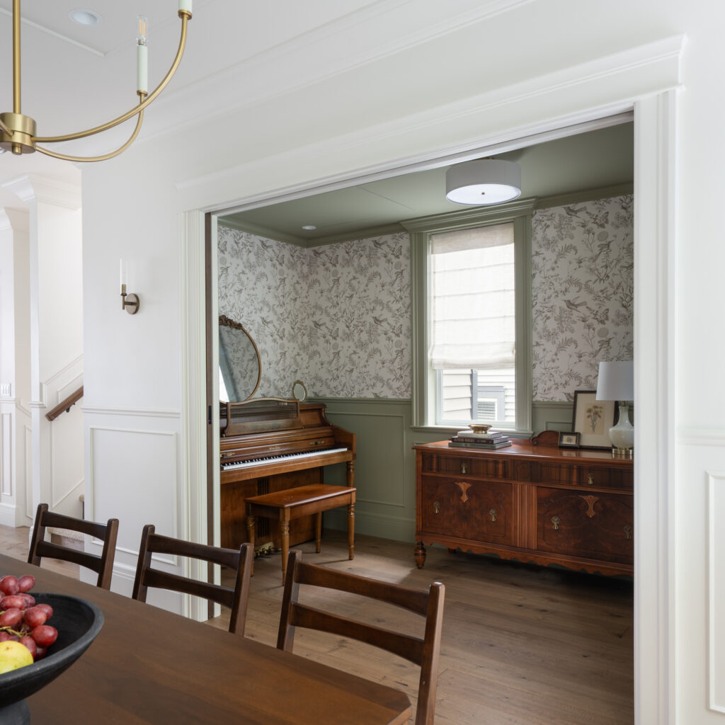 A dining area with a wooden table leads to a room with a piano, mirror, and sideboard. The room has floral wallpaper, green trim, a window with a shade, and wooden flooring.