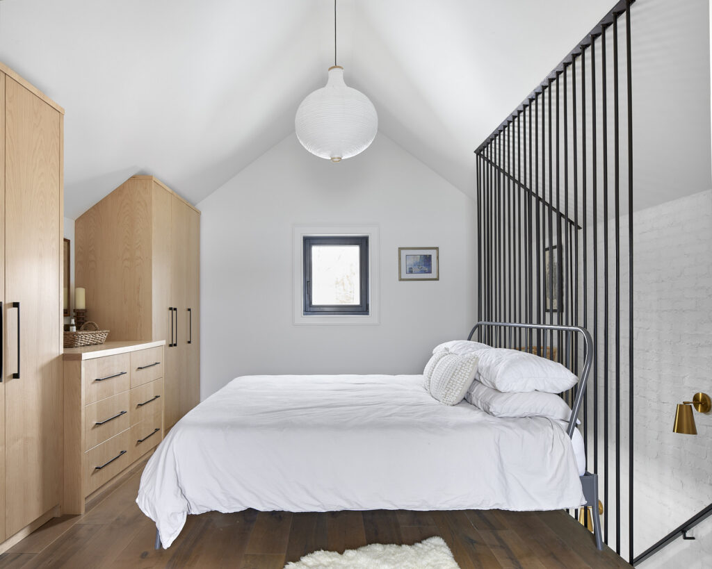 Minimalist bedroom with sloped ceiling, small window, light wood furniture, bed with white bedding, black metal divider, white brick wall, and a round hanging light fixture.