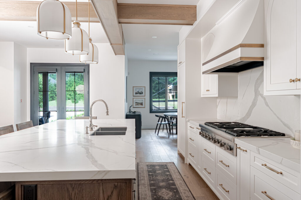 Modern kitchen with white marble countertops, pendant lights, white cabinets, gas stove, and a farmhouse sink. Large windows let in natural light; a dining area is visible in the background.