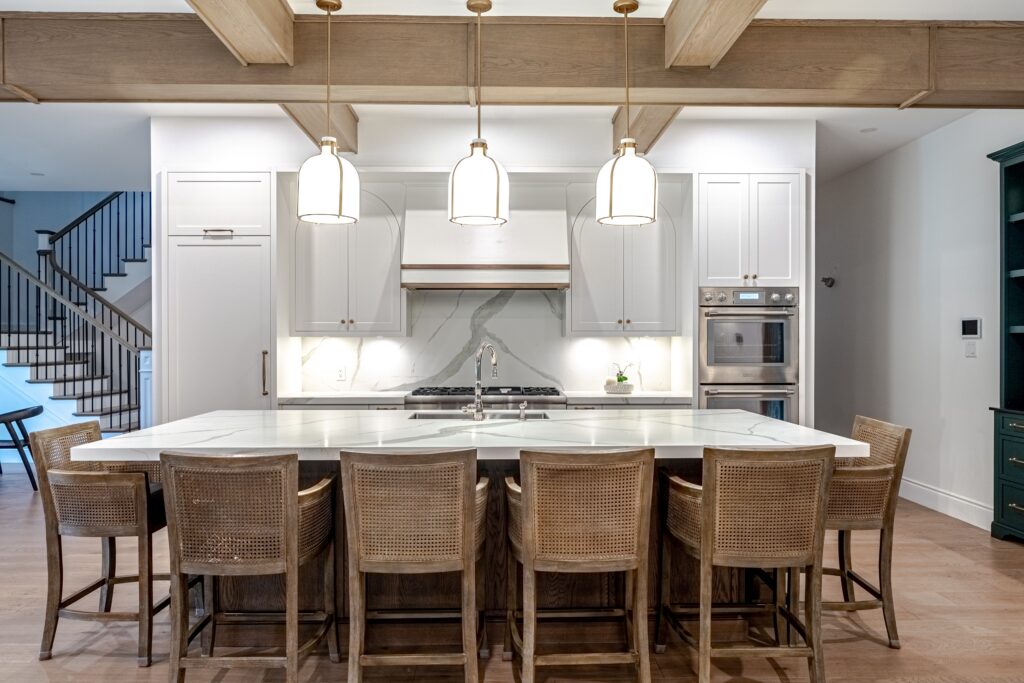 Modern kitchen with white cabinets, a marble island countertop, six wicker chairs, three pendant lights, built-in ovens, and a gas cooktop. Wooden beams and staircase are visible in the background.