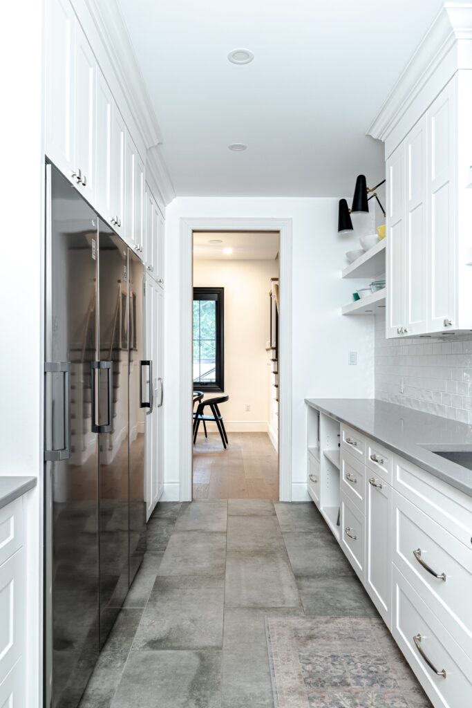 Modern white kitchen with stainless steel double fridge, white cabinets, gray countertops, and tiled floor. A doorway leads to a bright dining area with a window and dark chairs.