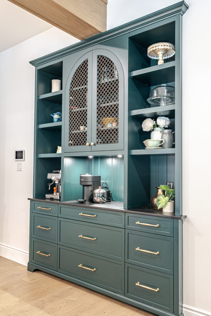 Green built-in cabinet with gold handles, open shelves displaying dishes and glassware, mesh doors at the top center, and a coffee machine on the counter beneath under-cabinet lighting.