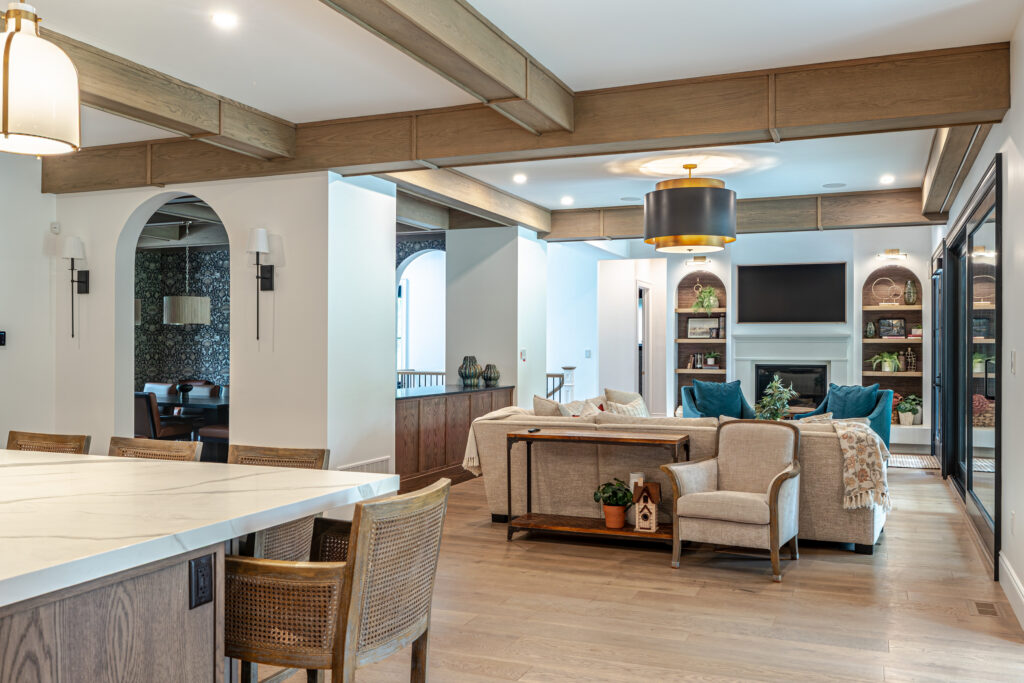 Open-concept living space with wood beams, kitchen island, dining chairs, cozy seating area, built-in shelves, fireplace, TV, and modern lighting. Light wood floors and neutral tones create a warm, inviting atmosphere.