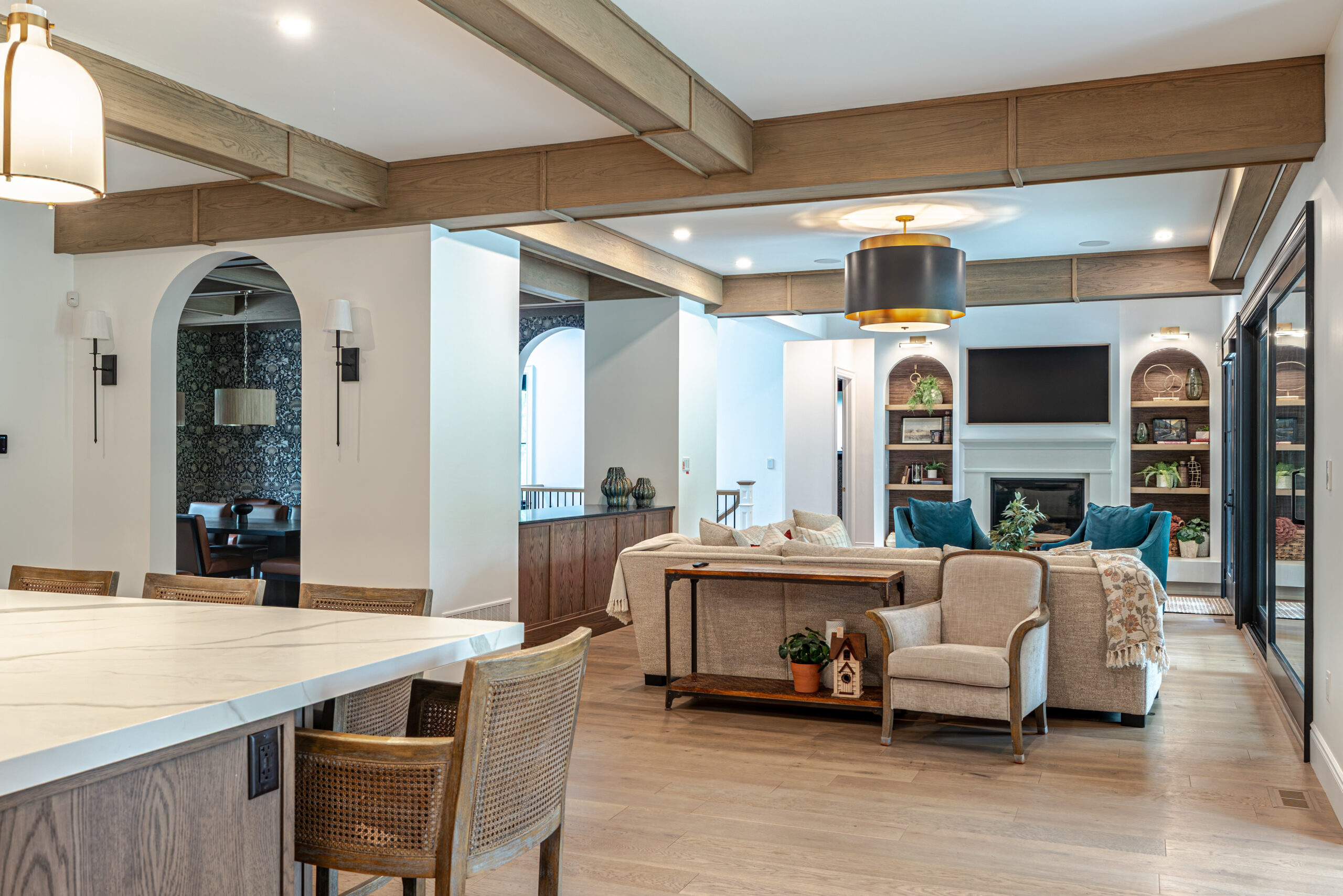Open-concept living space with wood beams, kitchen island, dining chairs, cozy seating area, built-in shelves, fireplace, TV, and modern lighting. Light wood floors and neutral tones create a warm, inviting atmosphere.