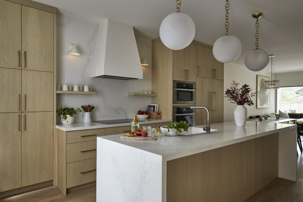 Modern kitchen with light wood cabinets, white countertops, globe pendant lights, built-in appliances, potted plants, and fresh produce on the island. A dining area is visible in the background.
