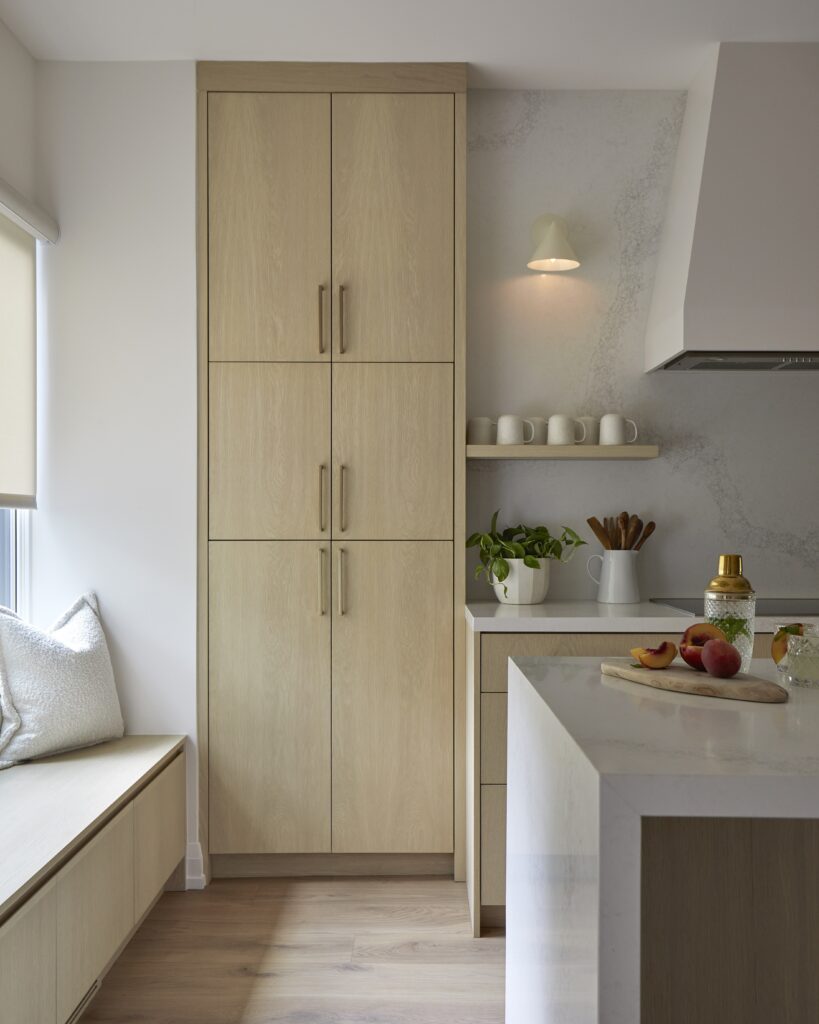A modern kitchen with light wood cabinets, a marble countertop, potted plants, mugs on a shelf, and fruit on the counter. A bench with cushions sits by a window, and a wall lamp is above the shelf.