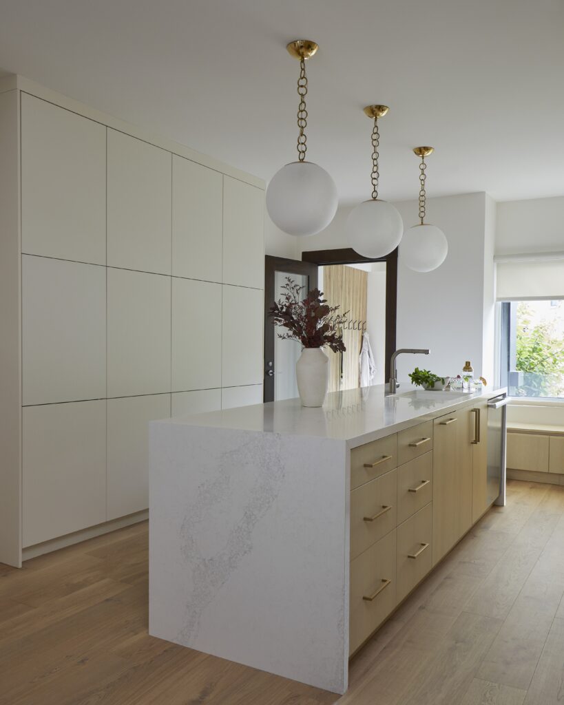 Modern kitchen with light wood floors, a large white island with gold handles, three round pendant lights, built-in cabinets, and a vase with dried flowers on the counter. Natural light streams in from a window.