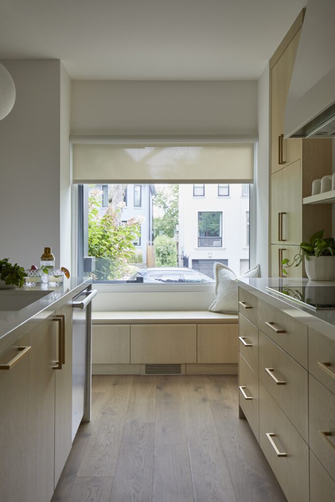 Modern kitchen with light wood cabinets, gold handles, and a built-in window bench with a cushion. A large window overlooks houses and trees outside, filling the room with natural light.