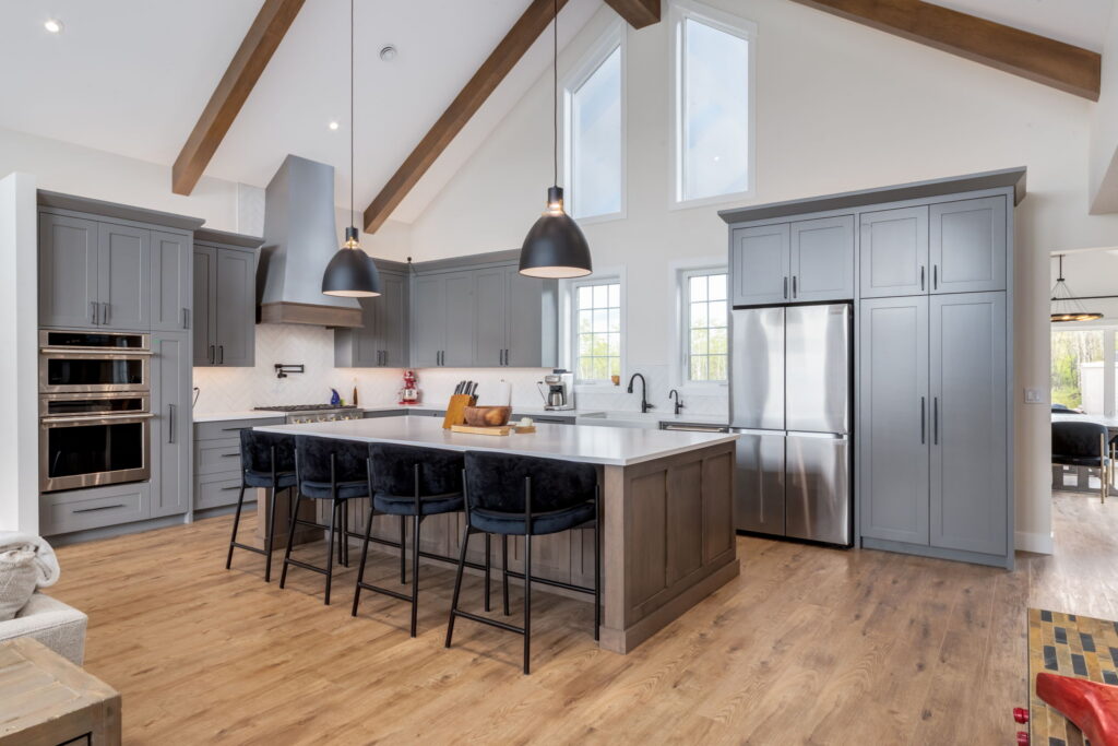 Modern kitchen with gray cabinets, stainless steel appliances, a large island with four black barstools, pendant lights, wood floor, and vaulted ceiling with wooden beams. Bright natural light comes through the windows.