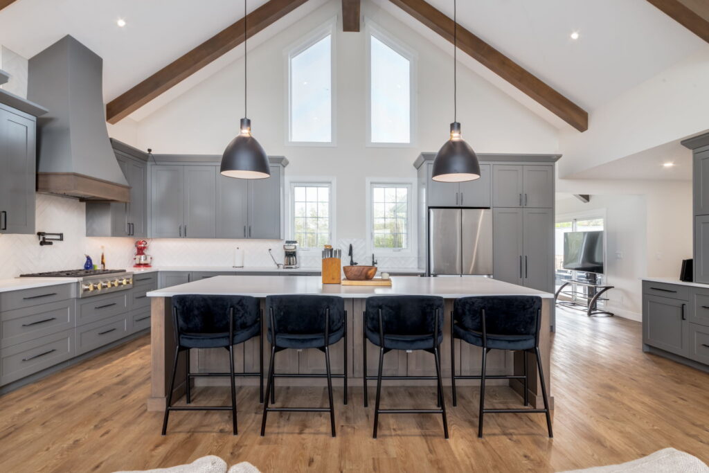 Modern kitchen with gray cabinets, large island with four dark barstools, pendant lights, stainless steel appliances, and wooden beams on the ceiling. Natural light comes through tall windows above the island.