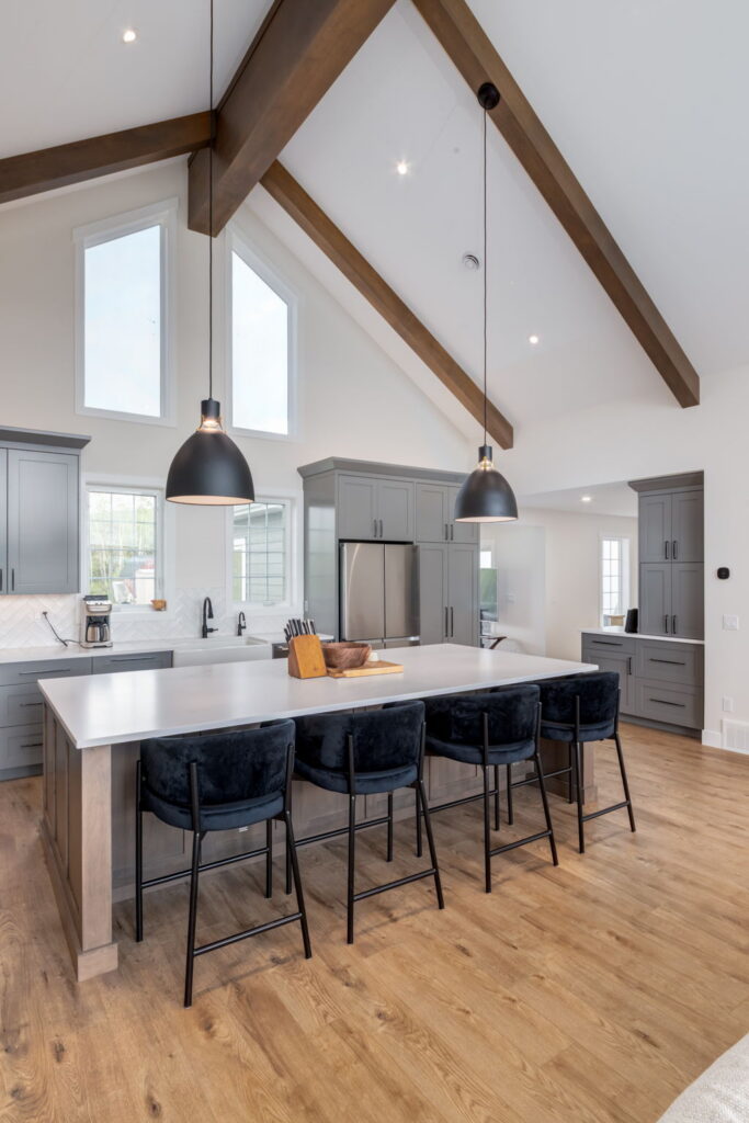 Modern kitchen with vaulted ceiling, exposed wood beams, gray cabinets, large island with white countertop, four black velvet barstools, pendant lights, and light wood flooring. Natural light streams through tall windows.