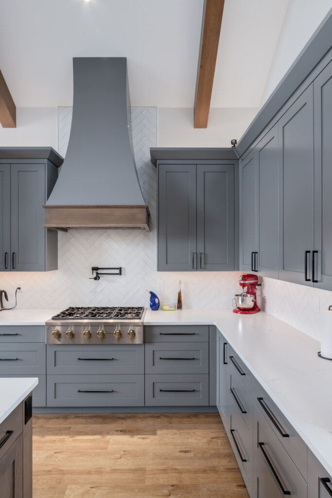 Modern kitchen with gray cabinets, white countertops, a stainless steel stove and range hood, wood flooring, and a red stand mixer on the counter. White herringbone backsplash and ceiling beams are visible.