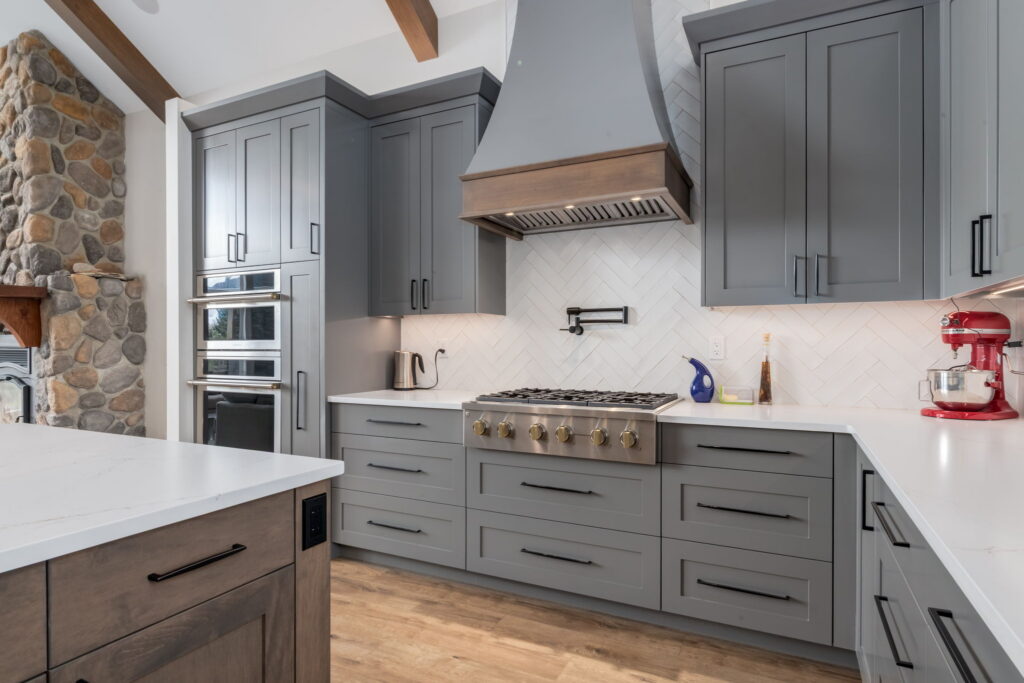 Modern kitchen with gray cabinets, stainless steel appliances, a built-in oven, gas stove, red stand mixer, and stone fireplace. White countertops and herringbone backsplash complete the look.
