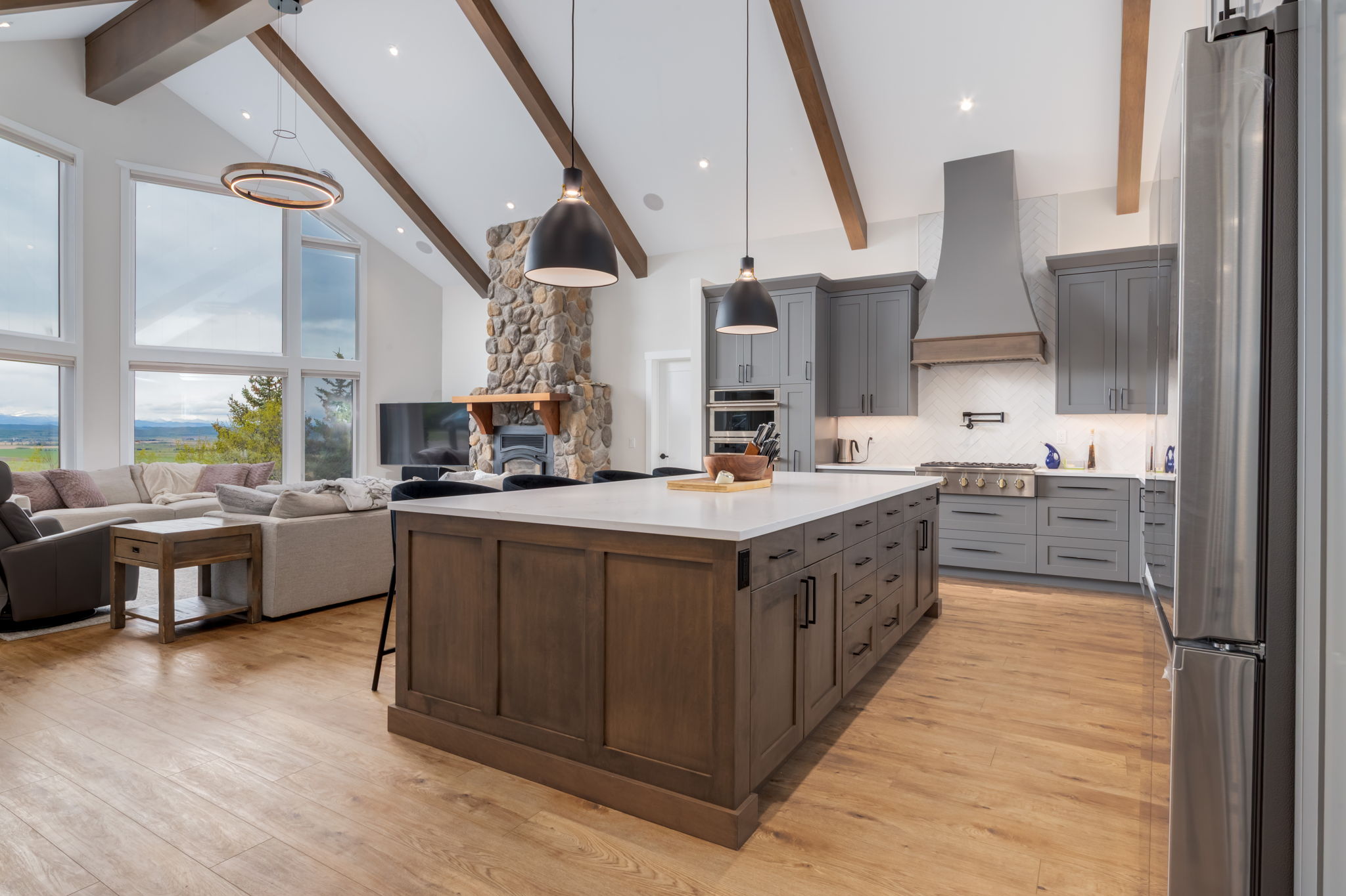 Modern kitchen with a large island, gray cabinets, pendant lights, stainless steel appliances, wood floors, and vaulted ceiling with beams. Living area with stone fireplace and large windows in the background.
