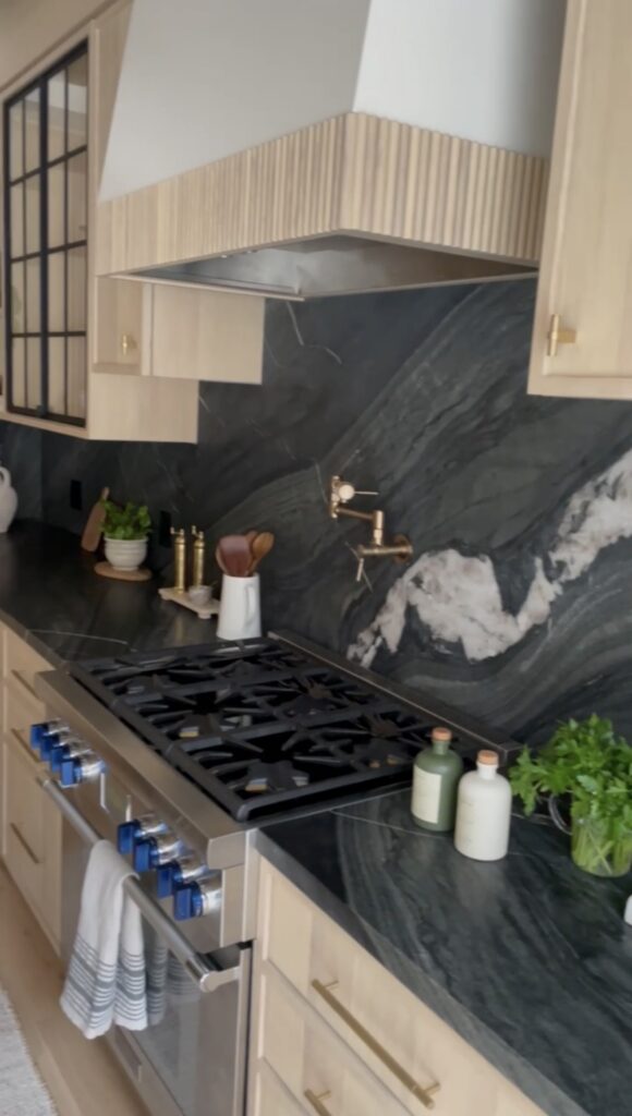 Modern kitchen with light wood cabinets, dark marble backsplash and countertops, stainless steel stove with blue knobs, labeled bottles, potted herbs, and cooking utensils displayed neatly on the counter.