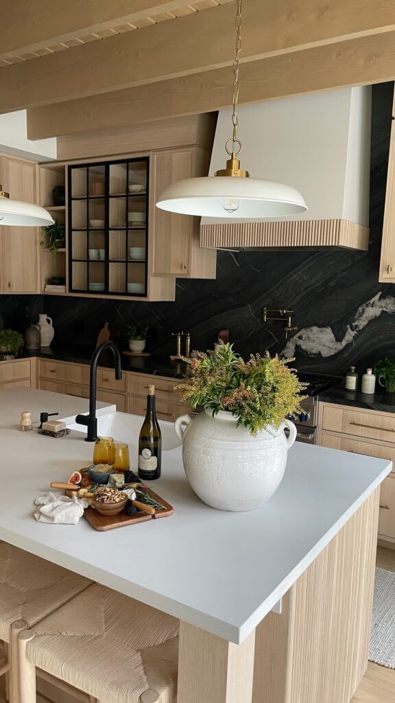 Modern kitchen with light wood cabinets, black marble backsplash, white island, a large white vase with flowers, a drink, a bottle, and a charcuterie board on the counter, plus pendant lights above.