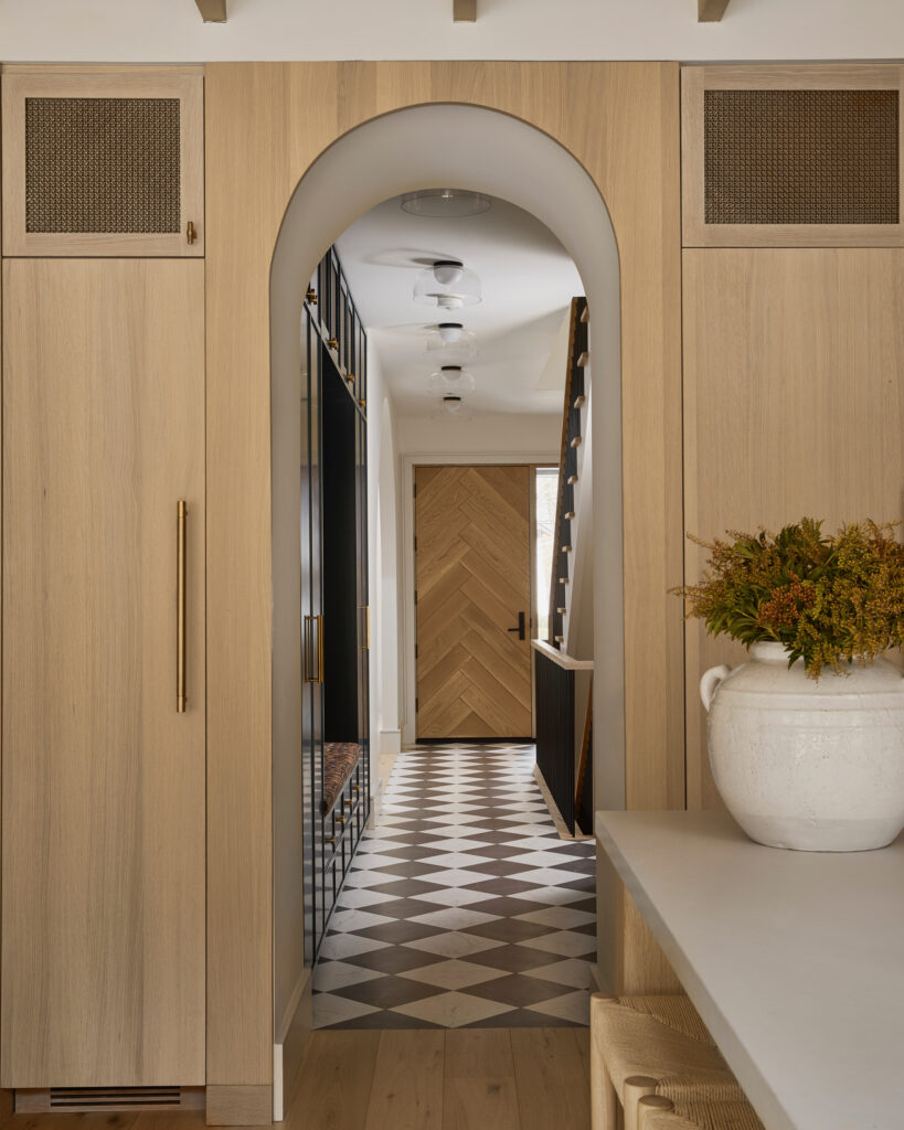 A hallway with light wood cabinets and an arched doorway leads to a chevron-patterned wooden front door. Black and white checkered floor tiles and a white vase with flowers are visible.