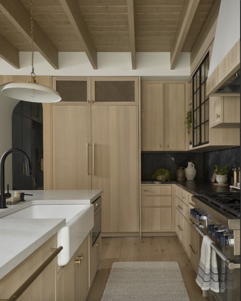 Modern kitchen with light wood cabinets, white countertops, a farmhouse sink, black fixtures, a gas stove, and a light rug. Exposed wood ceiling beams and dark backsplash add contrast. Decor items are on the counter.