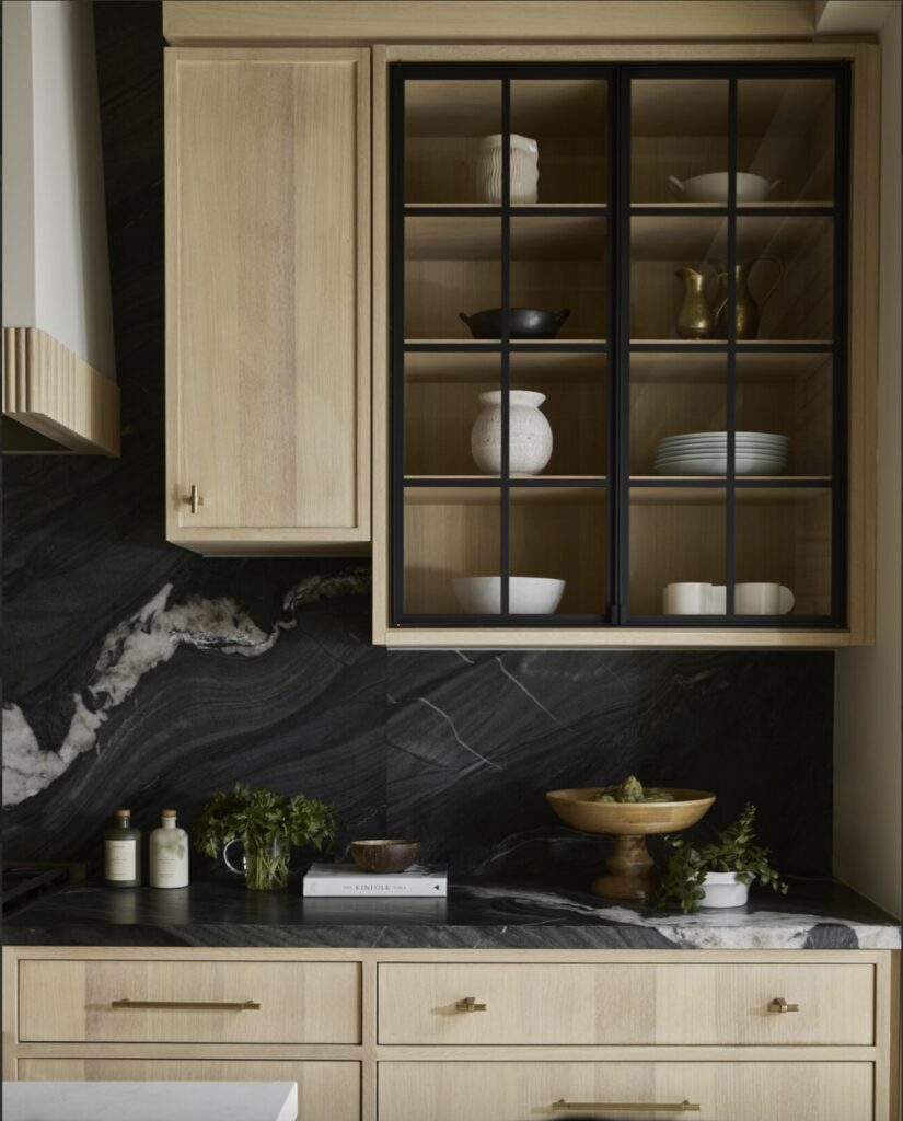 Light wood kitchen cabinets, black marble backsplash and countertop, glass-front upper cabinet with dishes, and a counter displaying a fruit bowl, plants, and bottles. Gold handles accent the drawers and doors.
