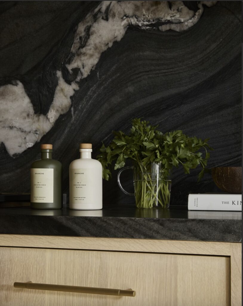 Two bottles, a clear glass cup with green leafy herbs, and a closed book sit on a dark marble countertop with dramatic white veining above a light wood drawer with a brass handle.