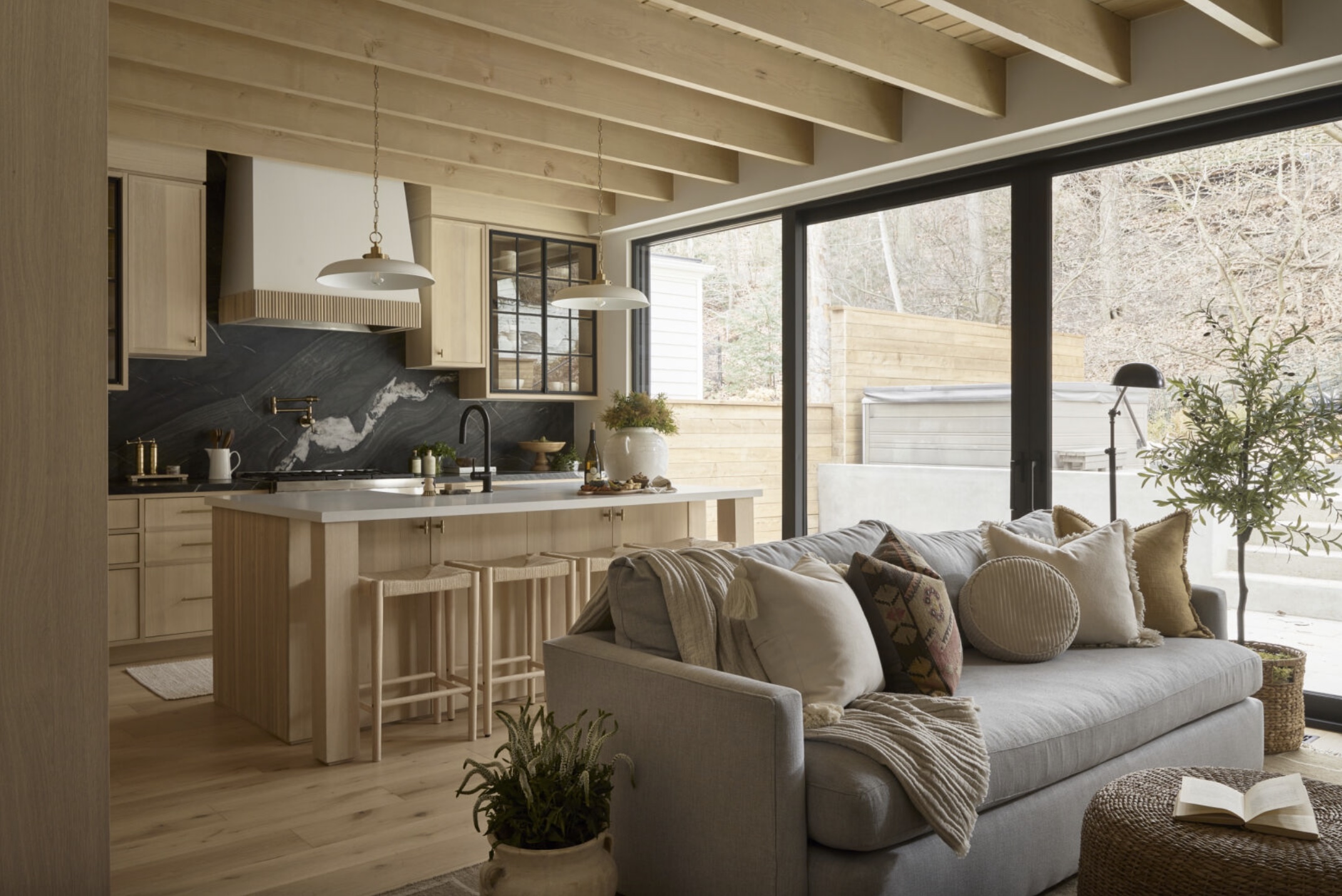 A cozy modern kitchen and living area with light wood, marble backsplash, island with stools, gray sofa, throw pillows, and large windows overlooking a fenced patio. Exposed wooden beams and plants add warmth.