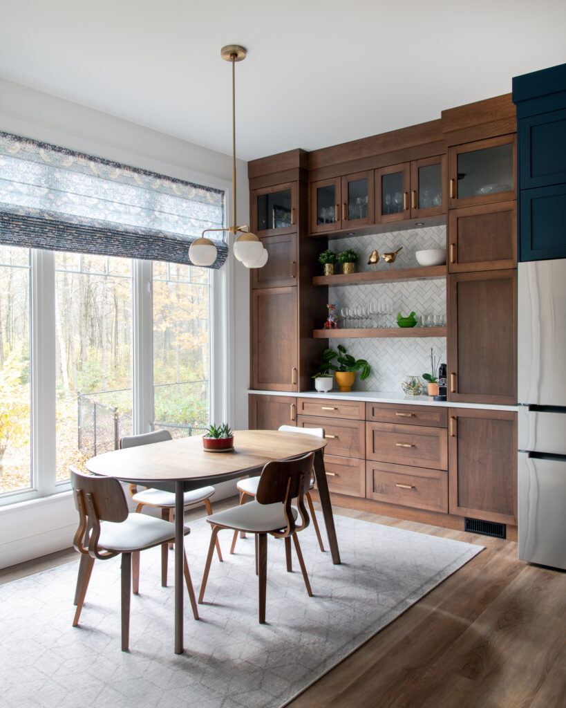 Modern kitchen with a wooden dining table and four chairs, large windows, wood cabinets with glass doors, open shelves with dishes, plants, and a light fixture above the table. Forest visible outside the windows.