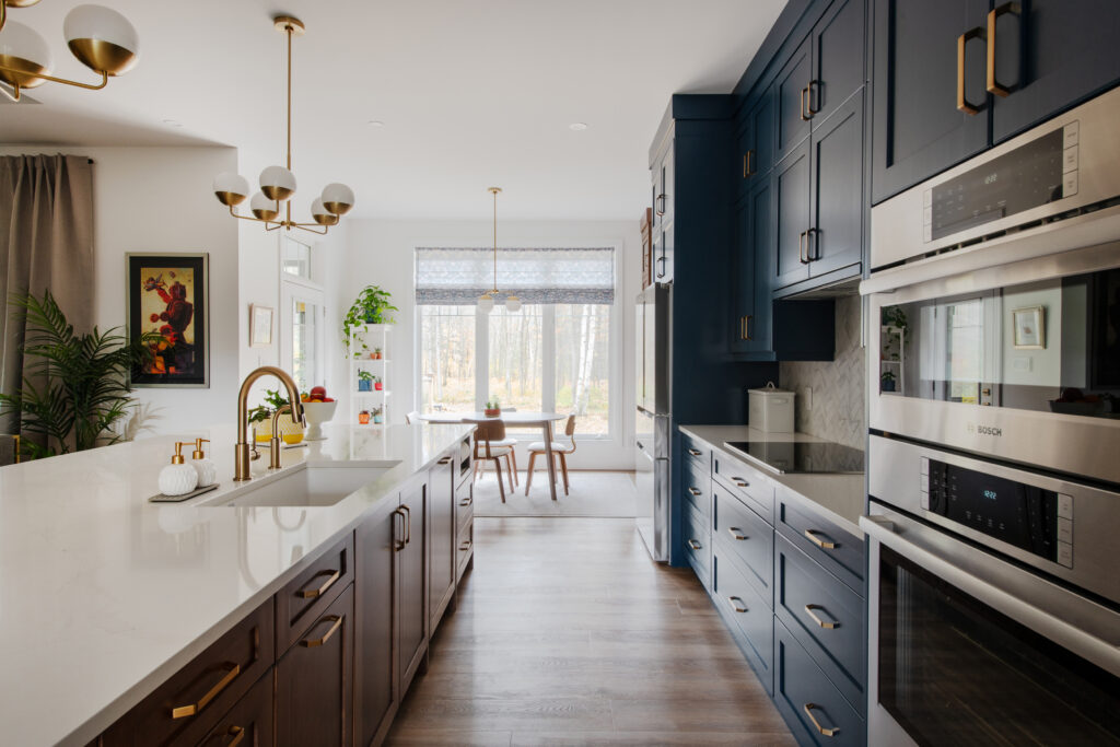 Modern kitchen with white countertops, navy and brown cabinets, double wall ovens, gold hardware, and a view into a bright dining area with large windows and wooden furniture.