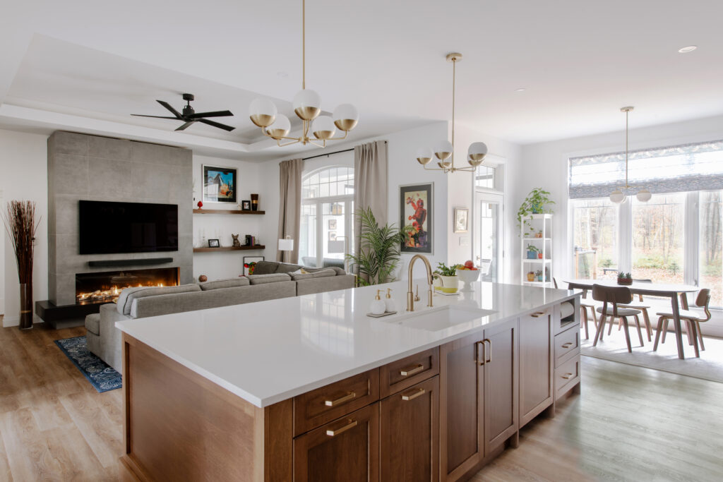 Modern open-concept kitchen and living room with a large island, wood cabinets, white countertops, grey sofa, fireplace, wall-mounted TV, dining area, big windows, and indoor plants.
