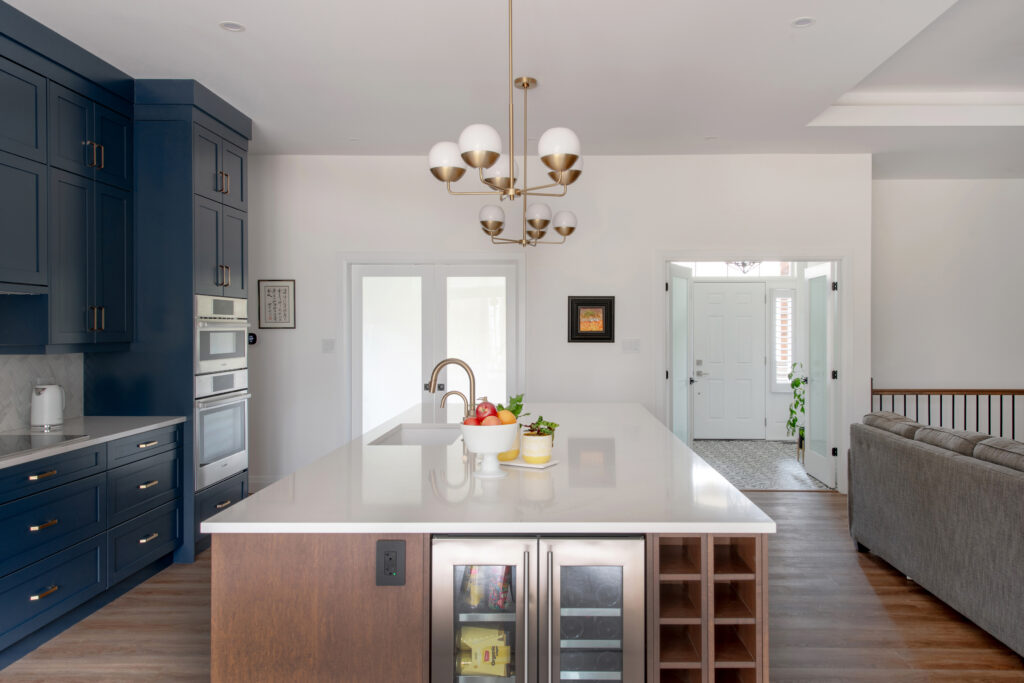 Modern kitchen with navy cabinets, a large white island, pendant chandelier, built-in oven, and glass door fridge. Living area and foyer visible in background with hardwood floors throughout. Bowl of fruit sits on the island.