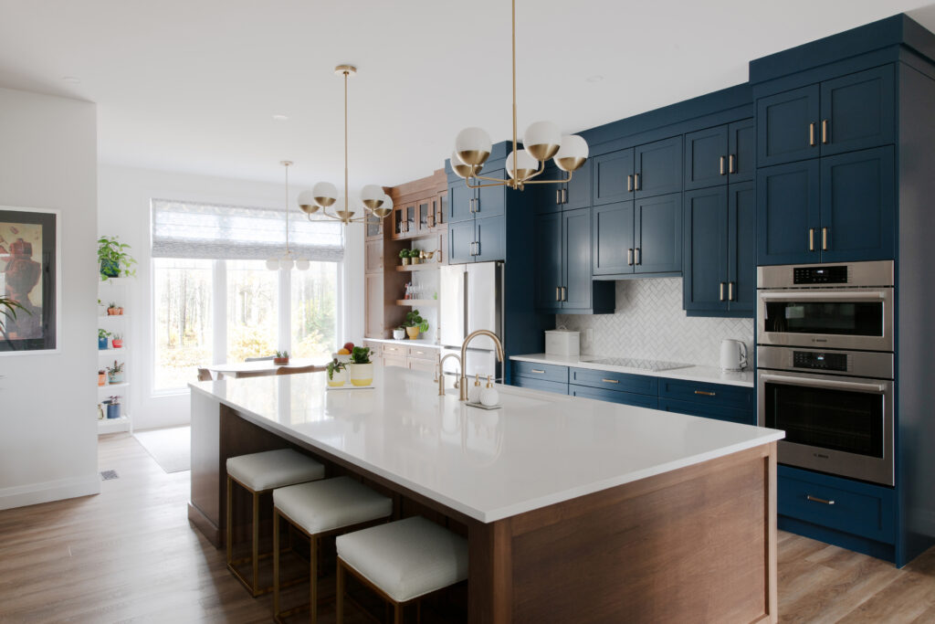 Modern kitchen with a large island, dark blue cabinets, gold fixtures, pendant lights, built-in ovens, and stools. Bright window lets in natural light. Wood floors and white countertops add a clean look.