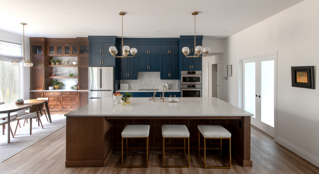 Modern kitchen with a large island, white countertop, navy cabinets, stainless steel appliances, gold light fixtures, and wooden flooring. Dining area with table and chairs is visible to the left.