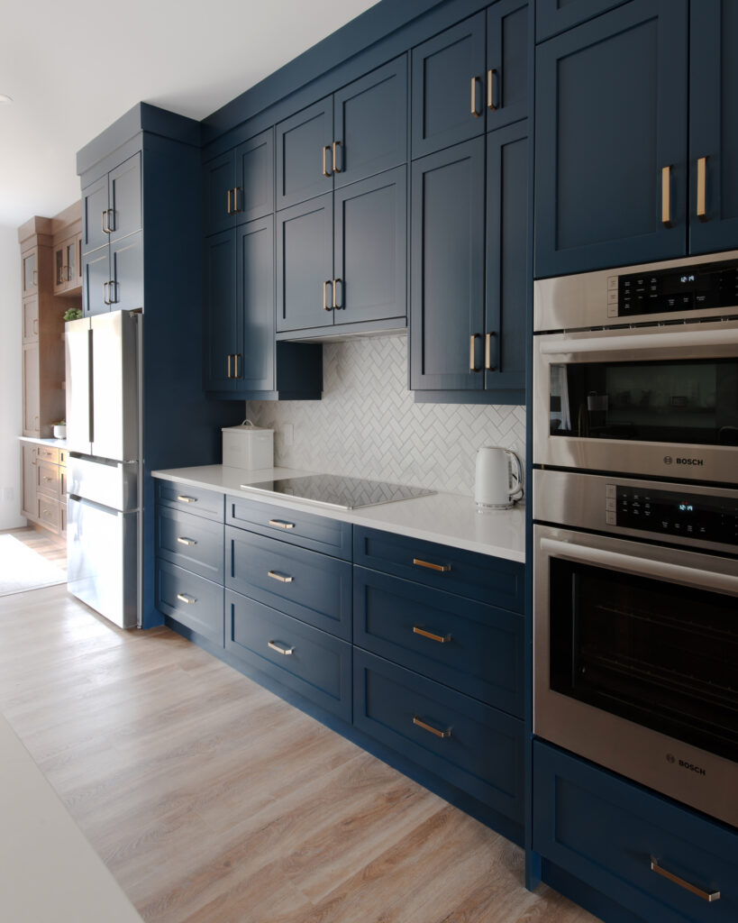 Modern kitchen with dark blue cabinets, stainless steel appliances, a white countertop, and herringbone tile backsplash. Light wood flooring adds warmth. The space is bright and organized.