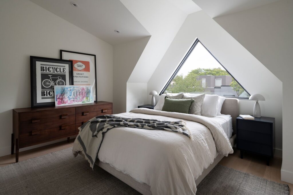 Modern bedroom with angled ceiling, triangular window, white bedding, green pillow, plaid blanket, dark wood dresser with framed art, two black nightstands, and two white lamps. Trees are visible outside the window.