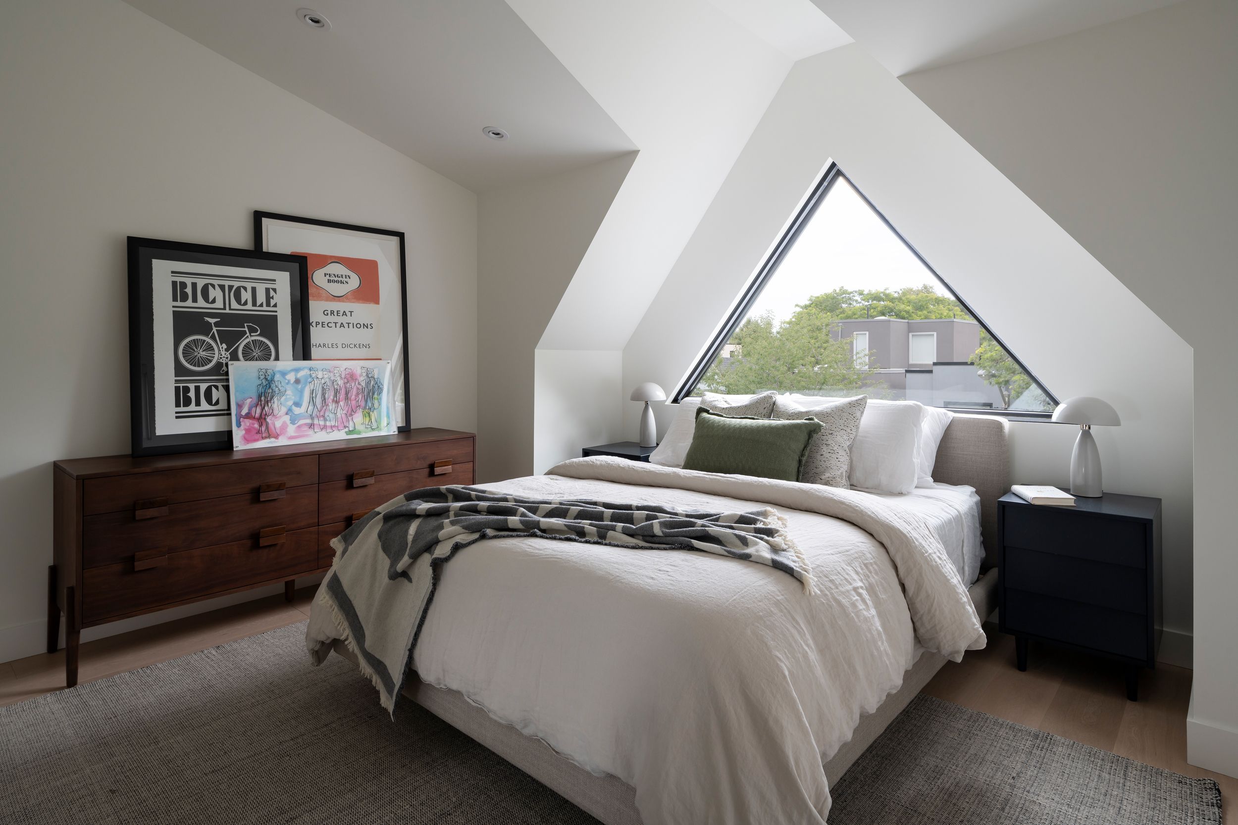 Cozy bedroom with a large triangular window, white bedding, a wooden dresser with framed art, black nightstands, and modern lamps. Natural light fills the minimalist space.