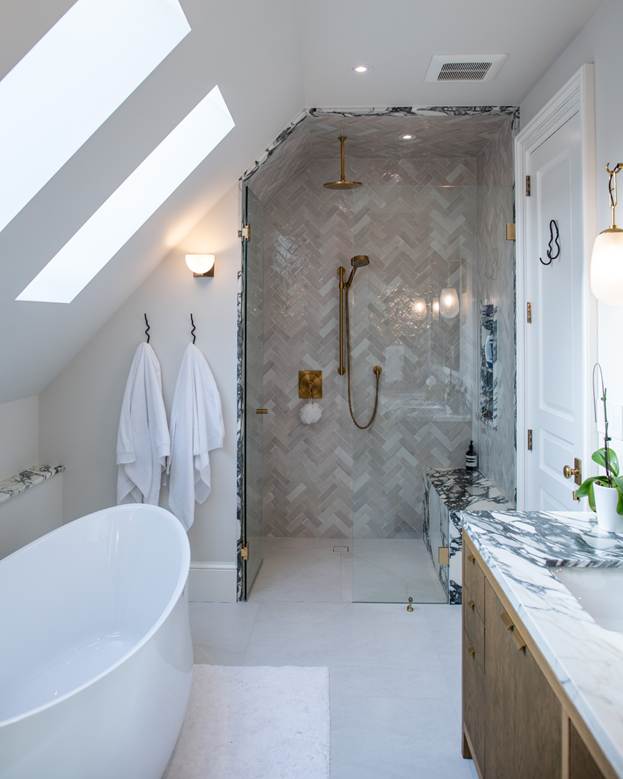 Modern bathroom with a freestanding bathtub, marble countertop, two white towels on hooks, and a glass-enclosed shower with gold fixtures and herringbone tile. Skylights provide natural light.