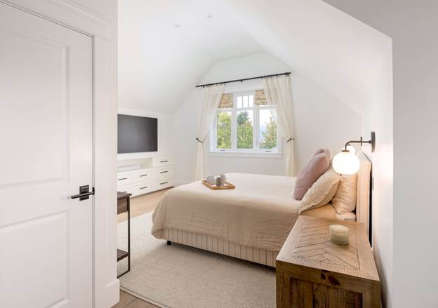 Bright bedroom with a beige bed, pink pillows, a wooden nightstand, wall lamp, large TV, dresser, and window with white curtains, letting in natural light and showing greenery outside.