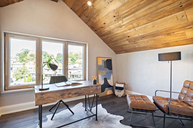 A cozy home office with a wooden desk, chair, and lamp by a large window, leather lounge chair, floor lamp, cowhide rug, and abstract art on the wall under a sloped wooden ceiling.