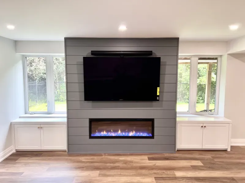 A modern living room with a wall-mounted TV above an electric fireplace, flanked by large windows and white built-in cabinets on each side.